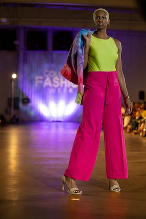 Model walks runway in bright yellow sleeveless top, hot pink wide-leg pants and denim jacket at fashion show.