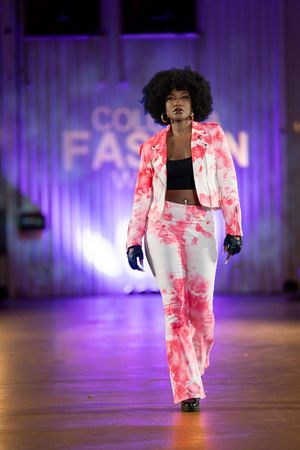 Model walks runway in pink and white tie-dye pantsuit during fashion show with purple lighting.