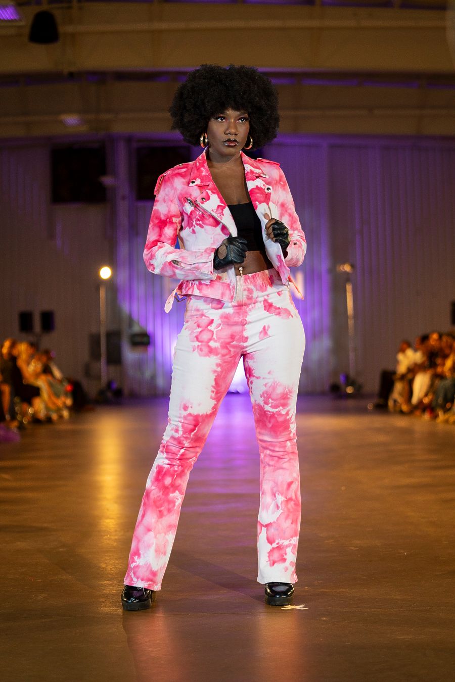 Model walks runway in vibrant pink and white tie-dye pantsuit during fashion show with purple lighting.