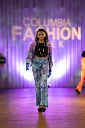 Model showcases tie-dye denim outfit on Columbia Fashion Week runway with purple backlit stage.