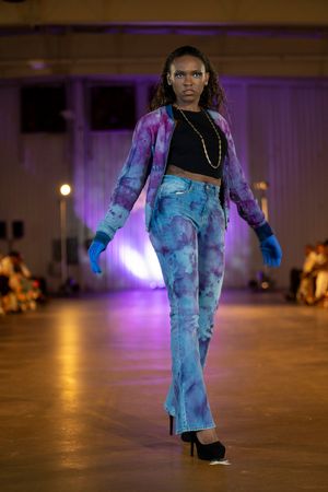 Model presents tie-dye denim ensemble with black crop top and gold chain on illuminated fashion runway.