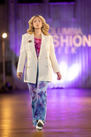 Fashion model walks runway in white blazer and blue tie-dye pants with purple lighting effects in background.