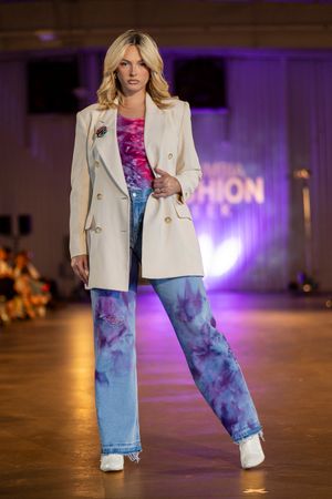 Fashion model walks runway wearing white blazer and tie-dye denim jeans at fashion show with purple lighting.