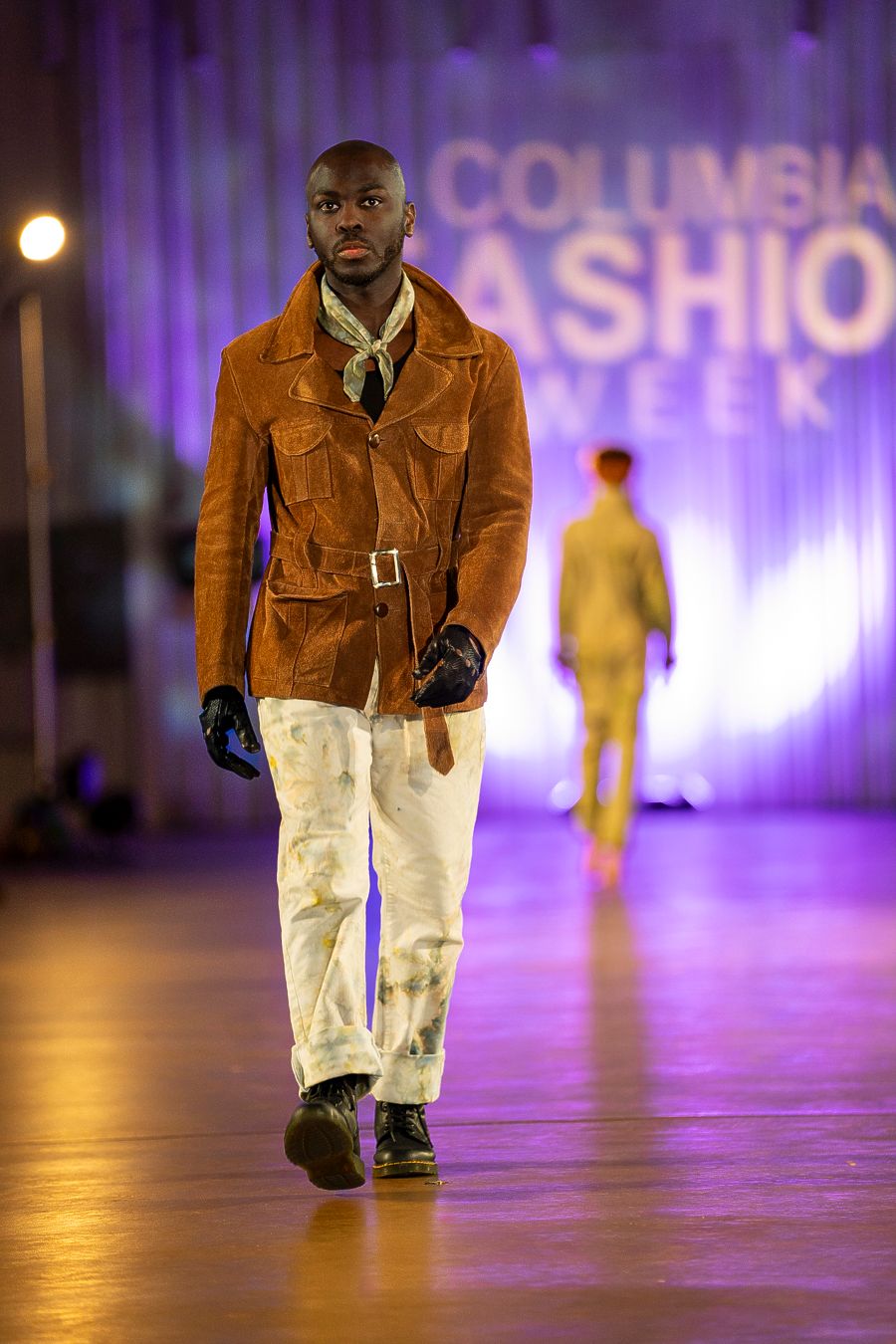 Model showcases brown suede jacket with white pants on illuminated runway during fashion event.