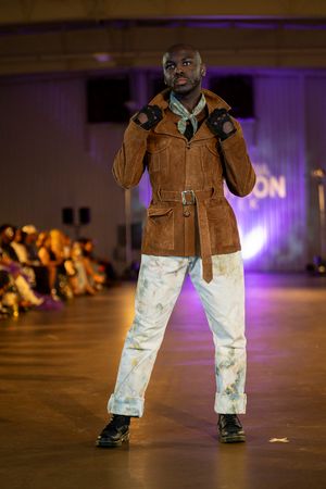 Close-up of model's lower body wearing brown jacket and distressed white pants on fashion runway.