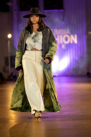 Model walks runway wearing white dress and camouflage military jacket at fashion show with purple lighting.