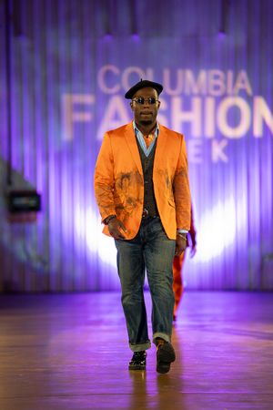 Model walks runway at Columbia Fashion Week wearing bright orange blazer, dark vest and jeans on purple-lit stage.