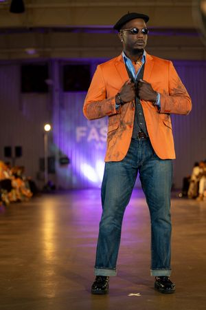Model walks runway wearing orange blazer and jeans during fashion show with purple lighting.