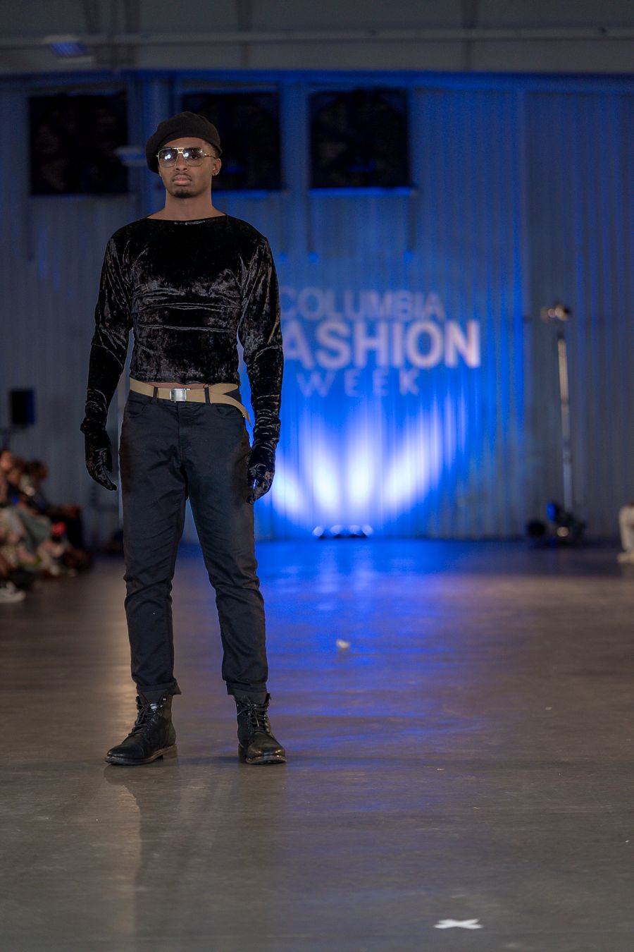 Model presents all-black streetwear ensemble on blue-lit runway during fashion show.