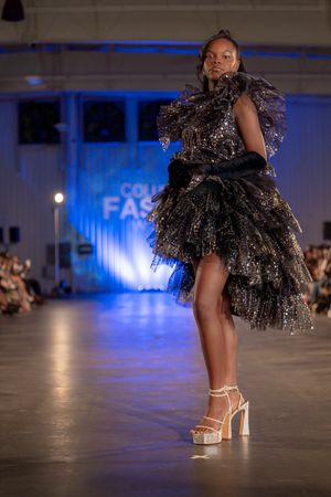 Model showcases dramatic black ruffled cocktail dress with silver platform sandals.