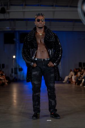 Model walks runway in edgy black textured ensemble with sunglasses.