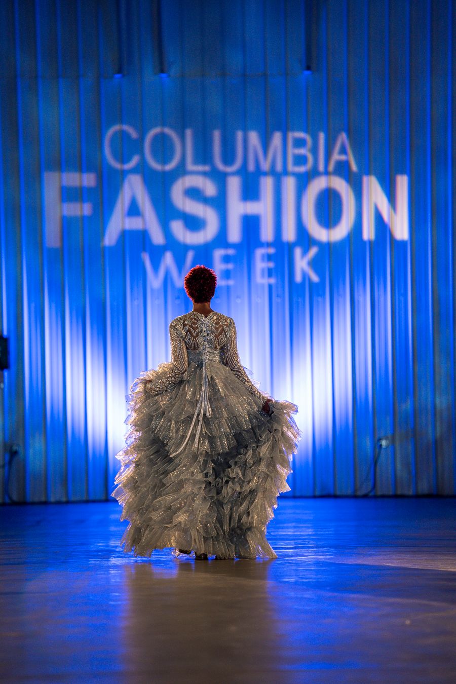 Silhouette of flowing ruffled gown against blue-lit Columbia Fashion Week backdrop.