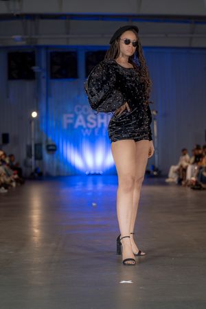 Model presents black velvet mini dress with strappy heels at fashion show.