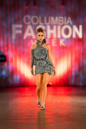 Fashion model walks runway in blue patterned top and nude bodysuit against red-lit backdrop at Columbia fashion show.