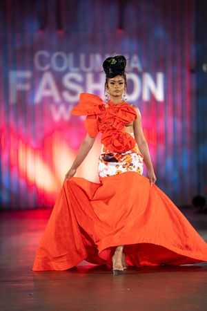Model walks runway in dramatic orange gown with ruffled details at Columbia Fashion Week show.