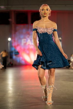 Model walks runway in navy blue off-shoulder cocktail dress with crystal embellishments and strappy heels at fashion show.