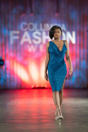 Model walks runway in blue dress at Columbia Fashion Week against vibrant pink and blue lighting.