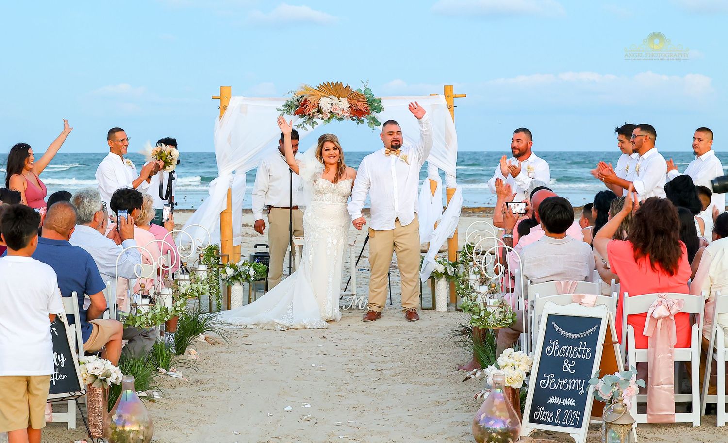 A beachfront wedding ceremony with white decor, floral arrangements lining the aisle, and guests seated on coral chairs.