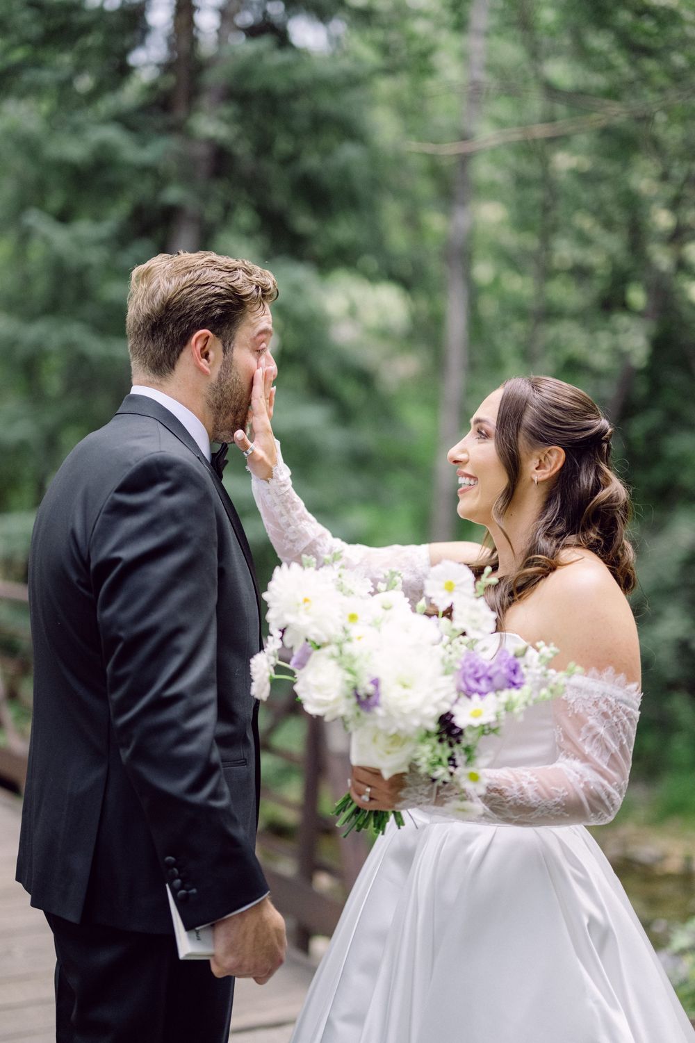 A bride and groom share a joyful moment in a forest setting, with the bride holding a bouquet of white and purple flowers.