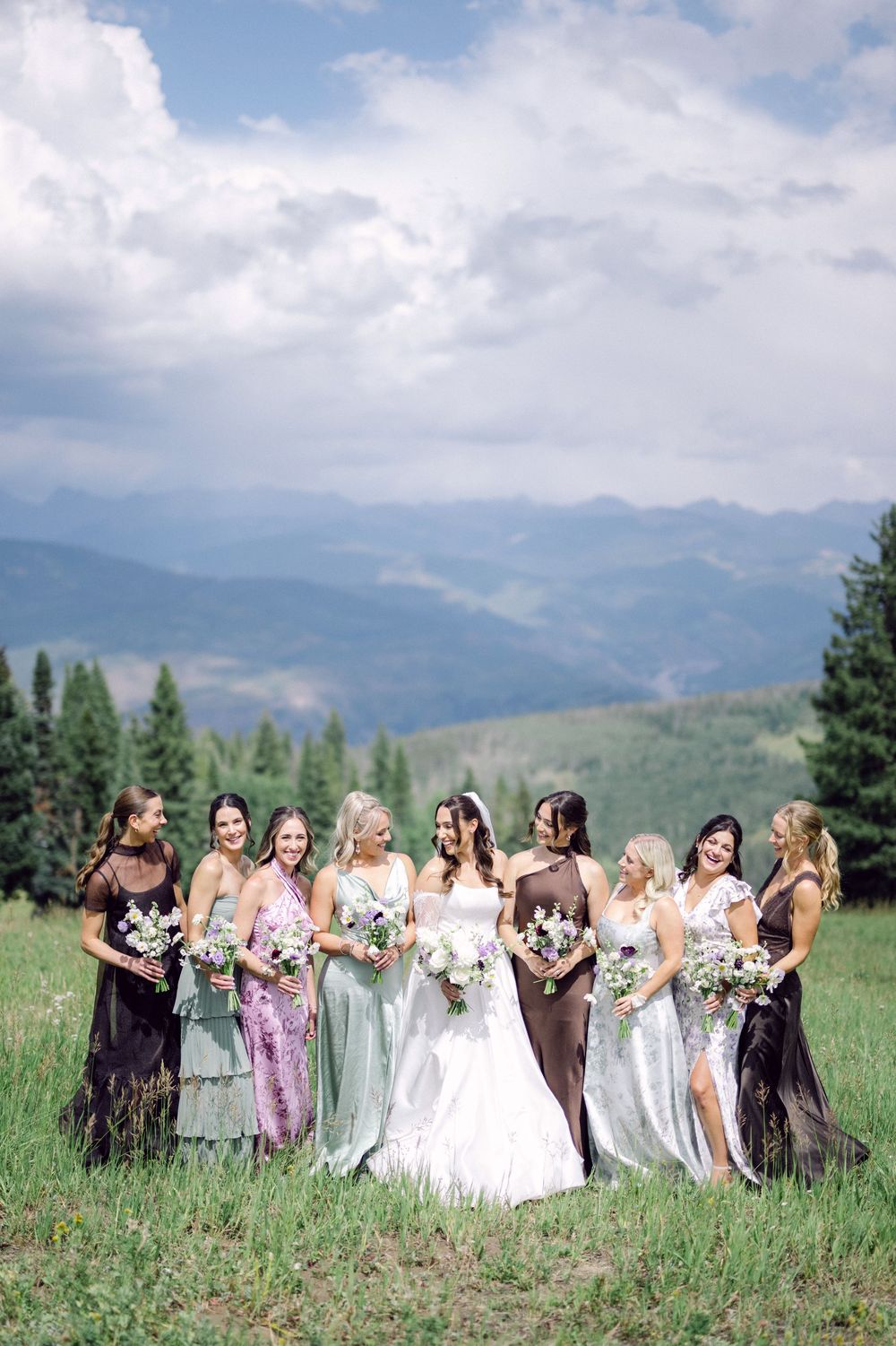 Bridal party in pastel dresses poses together in a scenic mountain field on a summer day.