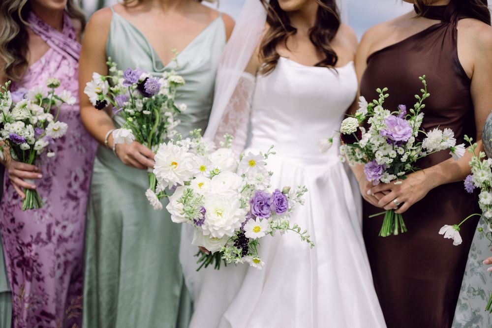 Bridesmaids in purple and sage dresses hold elegant white and purple floral bouquets.