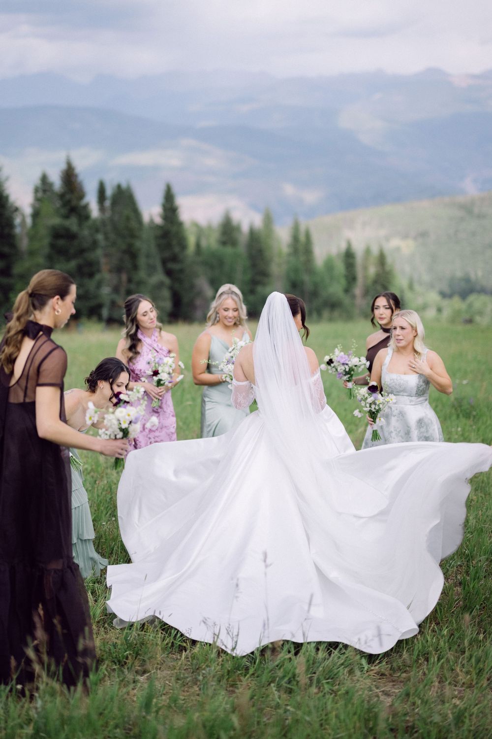 Bridesmaids in black dresses celebrate with bride in flowing white gown against mountain landscape backdrop.