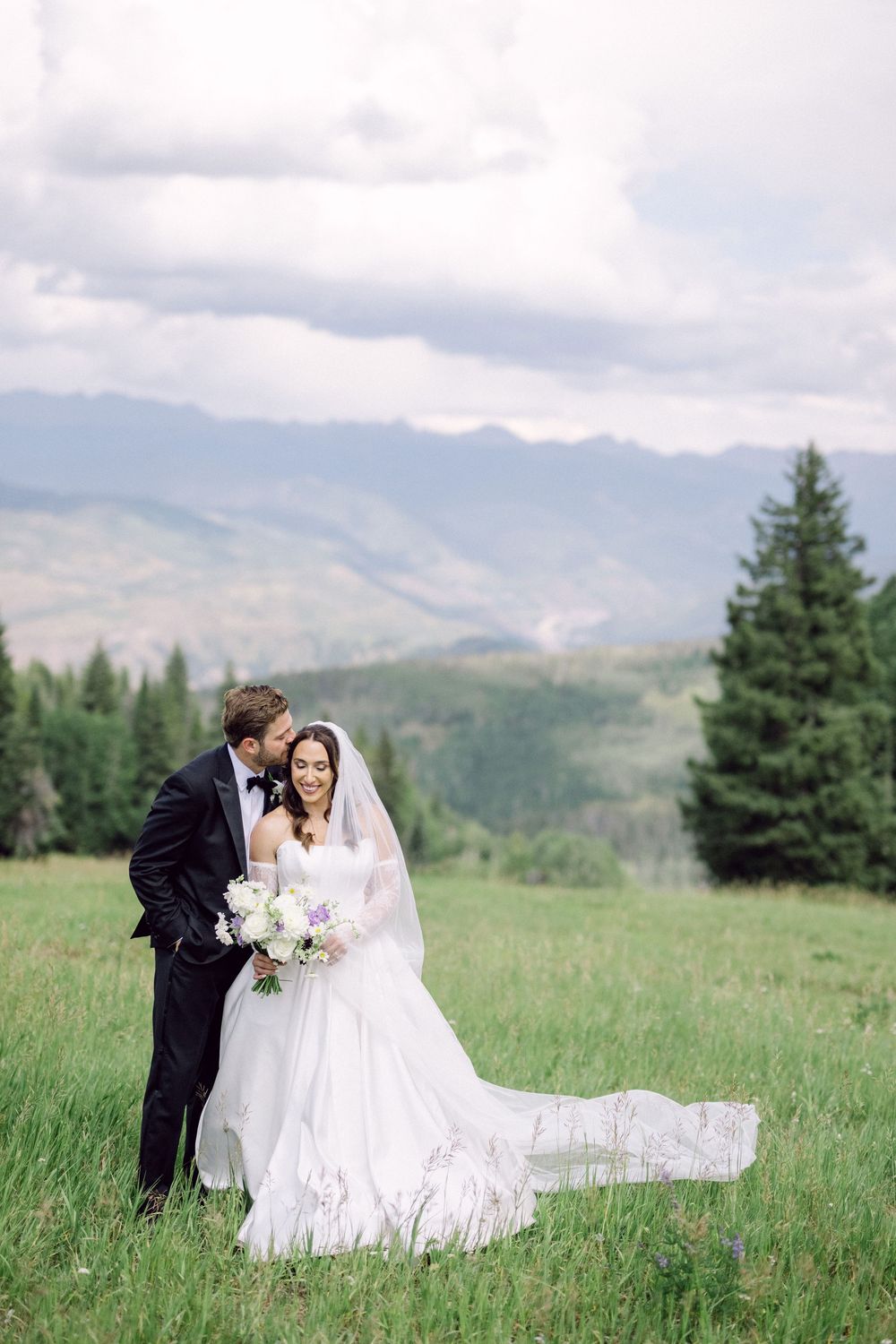 A couple in wedding attire embraces in a scenic mountain meadow with evergreen trees and cloudy skies in the background.
