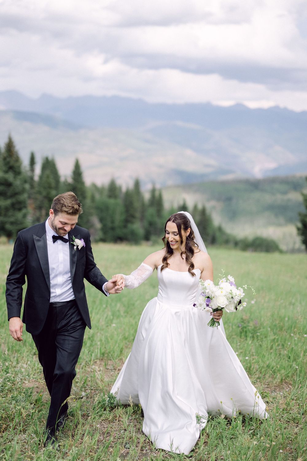 A bride and groom run playfully through a mountain meadow on their wedding day with evergreen trees in the distance.
