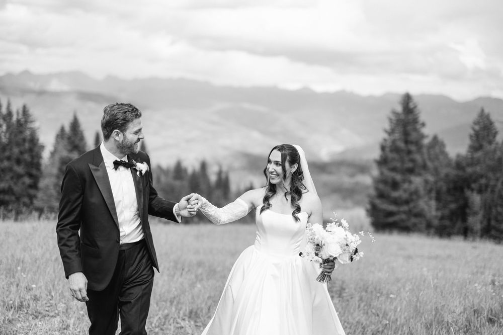 A couple in wedding attire shares a romantic moment in a scenic mountain meadow with evergreen trees in the background.