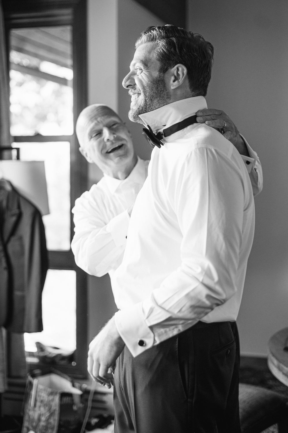 Two people share a candid laughing moment in black and white, wearing formal attire with suspenders and white shirts.