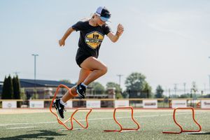 An athlete in a black t-shirt performs agility drills by jumping over orange hurdles on an outdoor sports field.