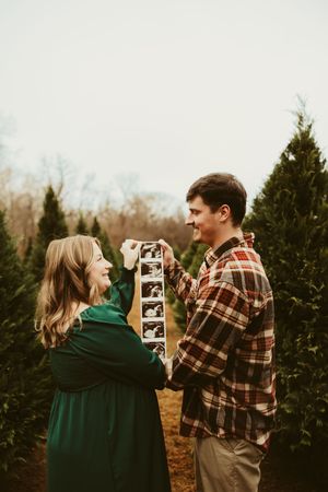 Expecting couple smiling at ultrasound photos at a Christmas tree farm during maternity session.
