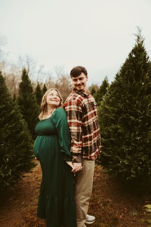 Pregnant woman in green dress and partner holding hands smiling at Christmas tree farm maternity shoot.