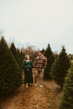 Expecting couple walking hand-in-hand through rows of evergreen trees during outdoor maternity session.