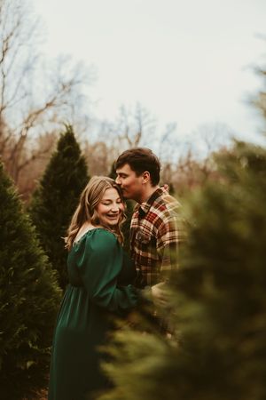 Man kissing laughing pregnant woman in green dress among Christmas trees during romantic maternity shoot.