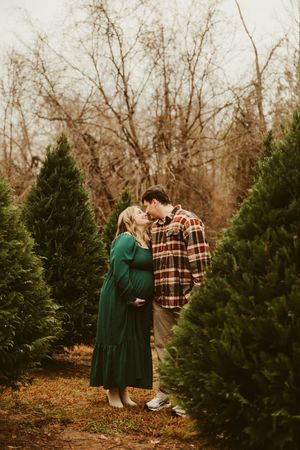 Pregnant couple sharing a tender kiss among evergreen trees at a Christmas tree farm.