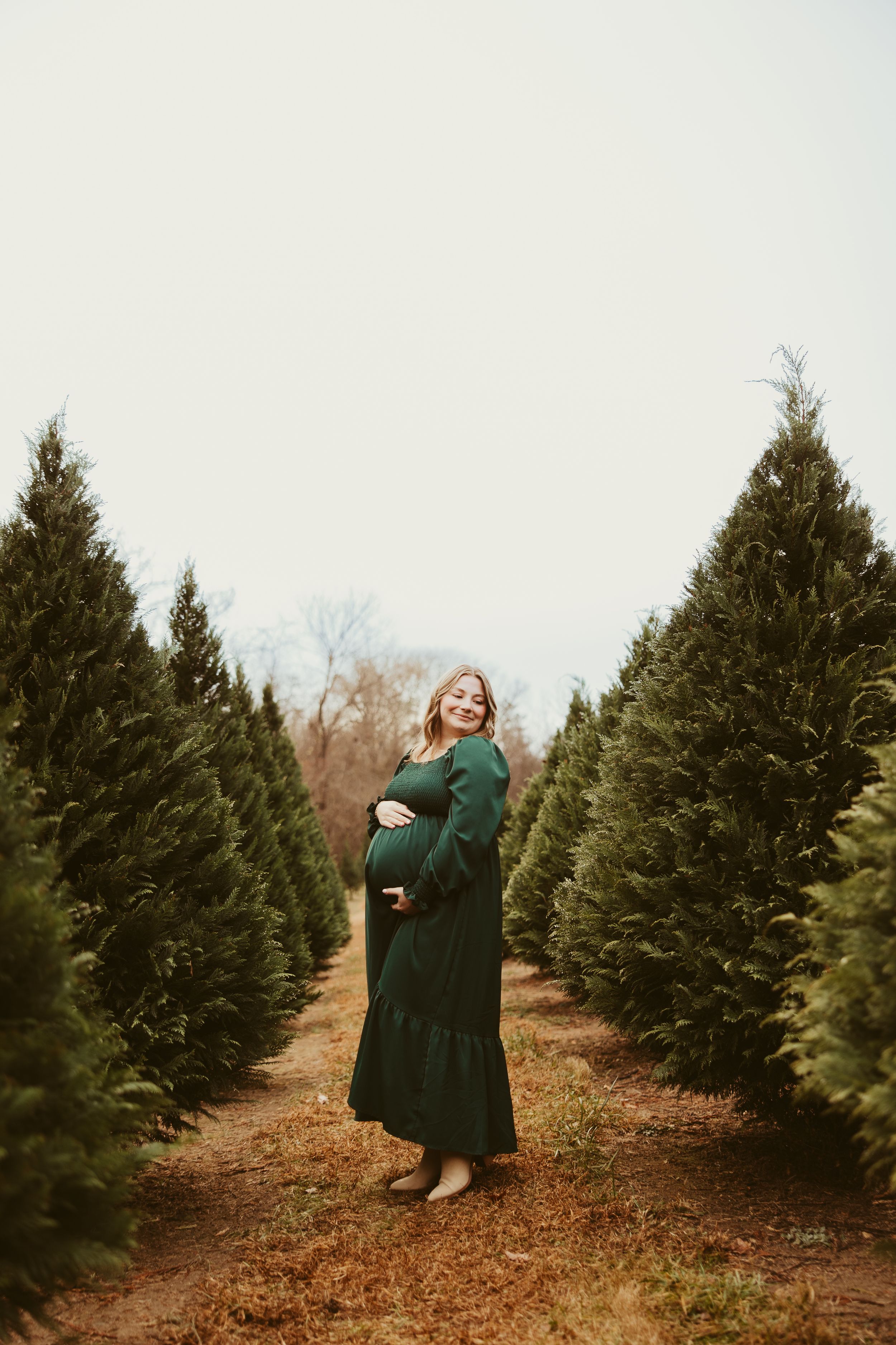 Smiling pregnant woman in a green dress cradling her baby bump at a Christmas tree farm.