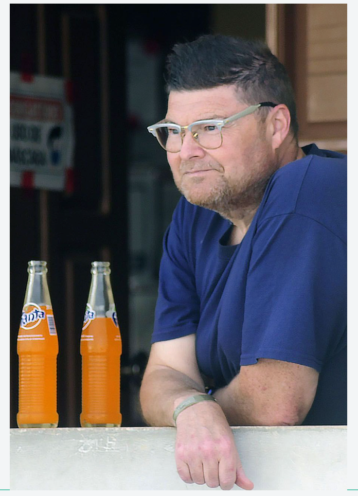 A person in a navy blue shirt sits at a table with two orange Fanta bottles, looking contemplatively at the beverages.