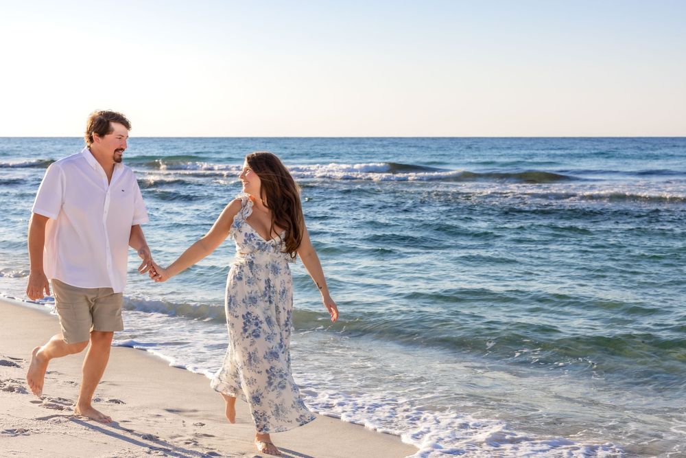 Couple holding hands and running playfully along a sunny beach with waves crashing on the shore.
