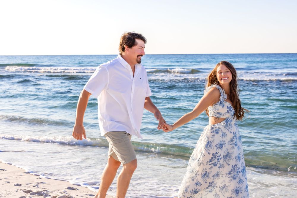 Couple in white outfits walking hand in hand along a sunny beach with ocean waves in the background.