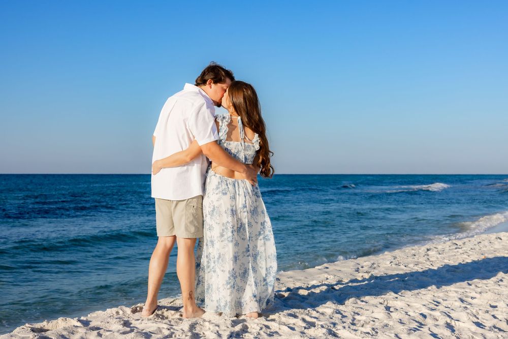 A romantic couple embraces on a white sandy beach with turquoise ocean waves and clear blue sky in the background.
