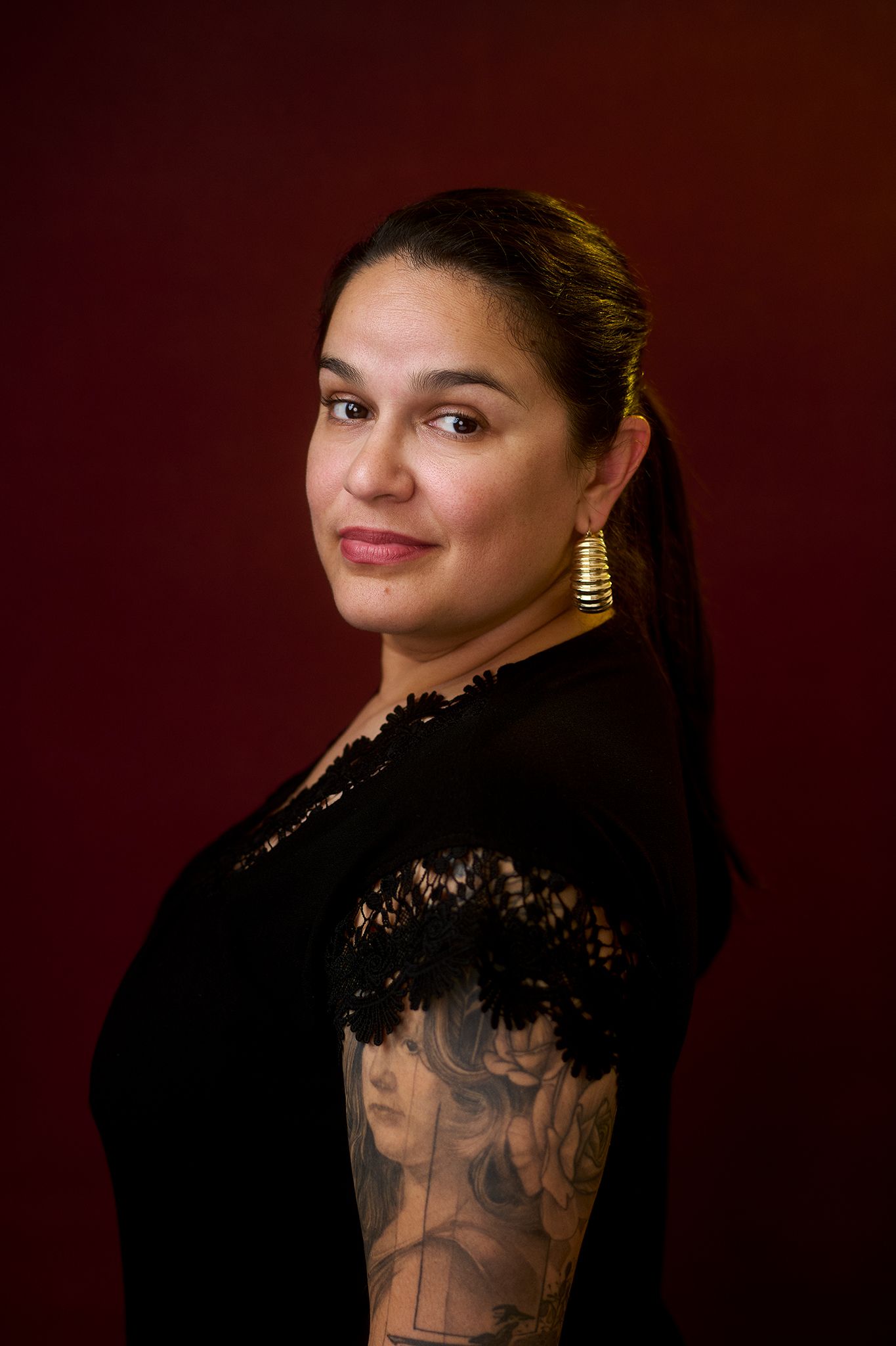 Woman in black lace top with floral arm tattoo and gold hoop earrings poses against dark red background.