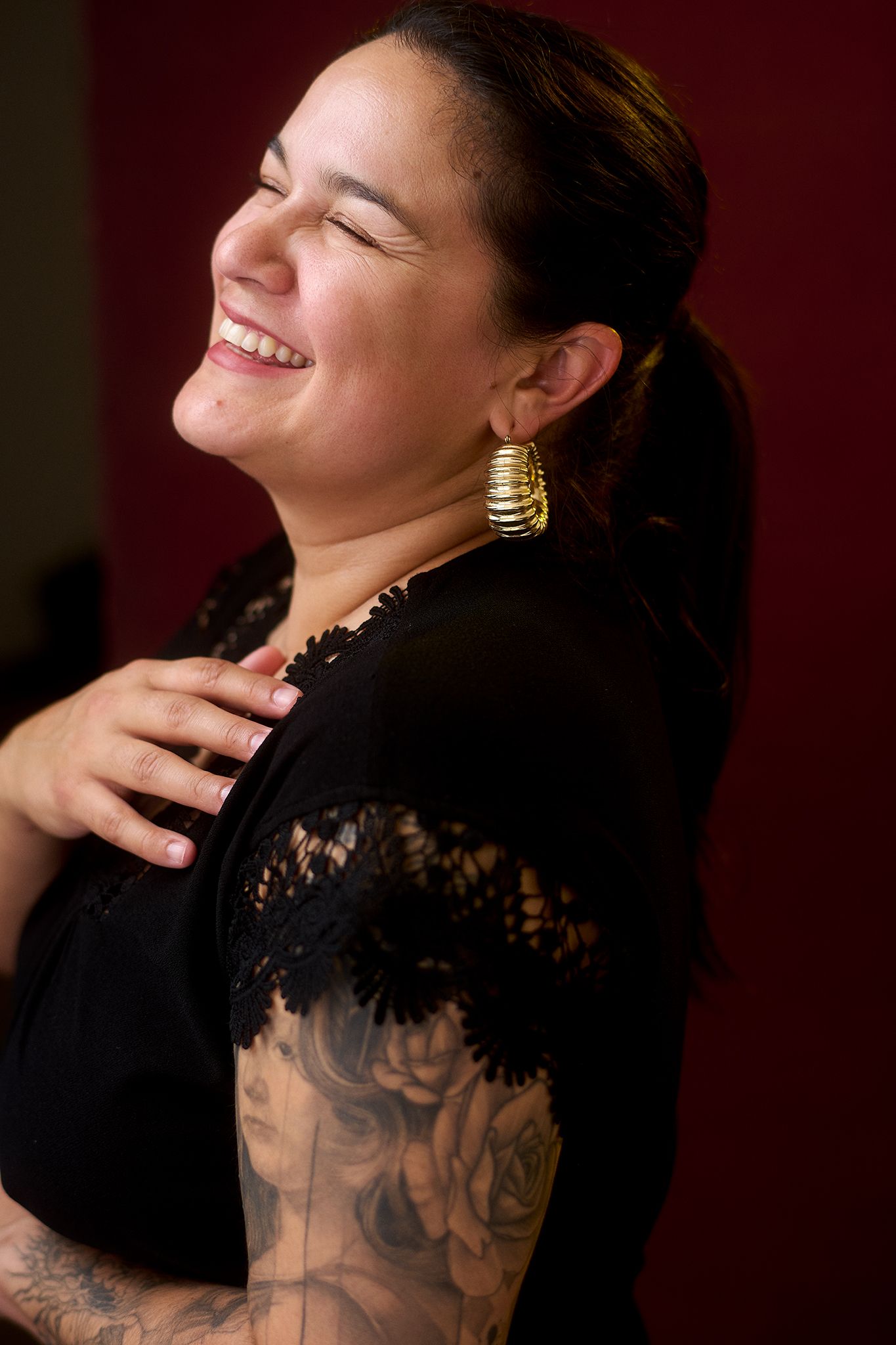 Laughing woman with tattoo sleeve, gold hoop earrings, and black lace top poses confidently against dark red background.