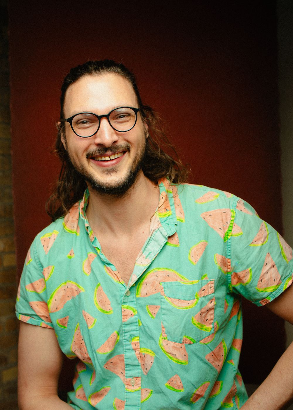 Smiling man with glasses and long hair wearing colorful watermelon print shirt against red background.