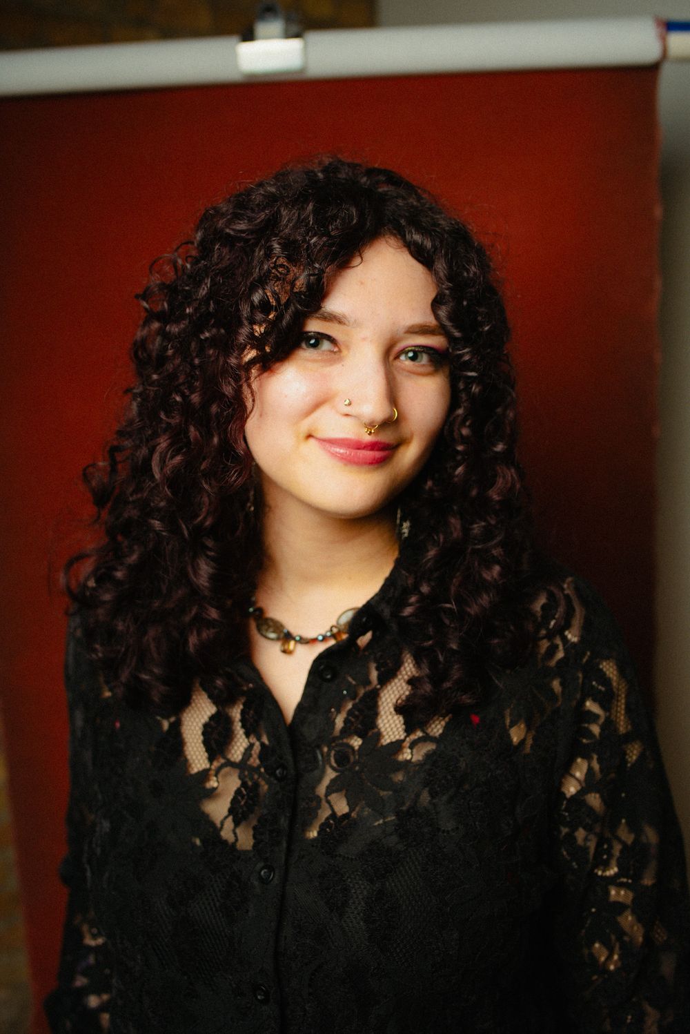 Young woman with voluminous dark curly hair wearing black lace blouse and nose ring smiling warmly.