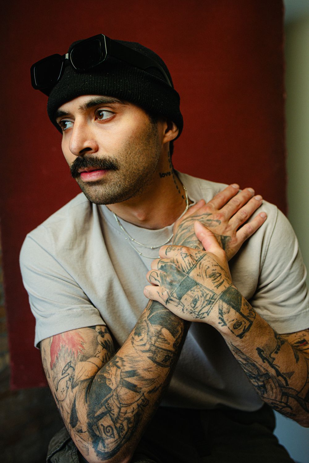 Tattooed man with mustache and black beanie wearing gray t-shirt posing against red wall.