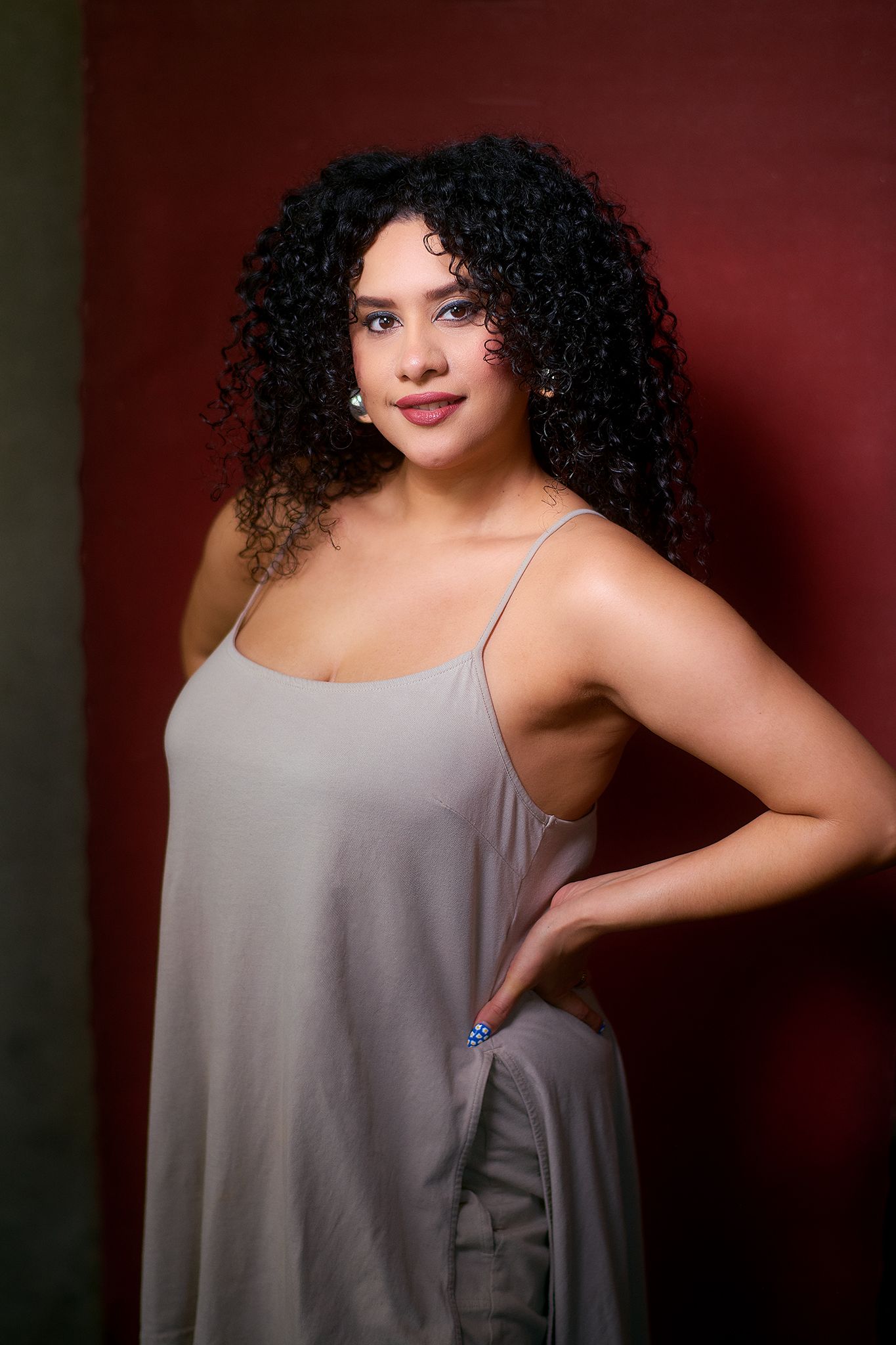 Young woman with curly black hair wearing a gray slip dress, posing confidently against a dark red background.