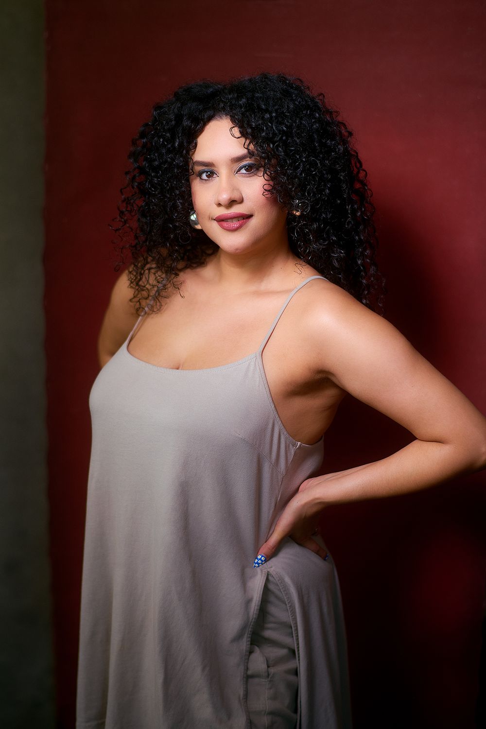 Young woman with curly black hair wearing a gray slip dress, posing confidently against a dark red background.