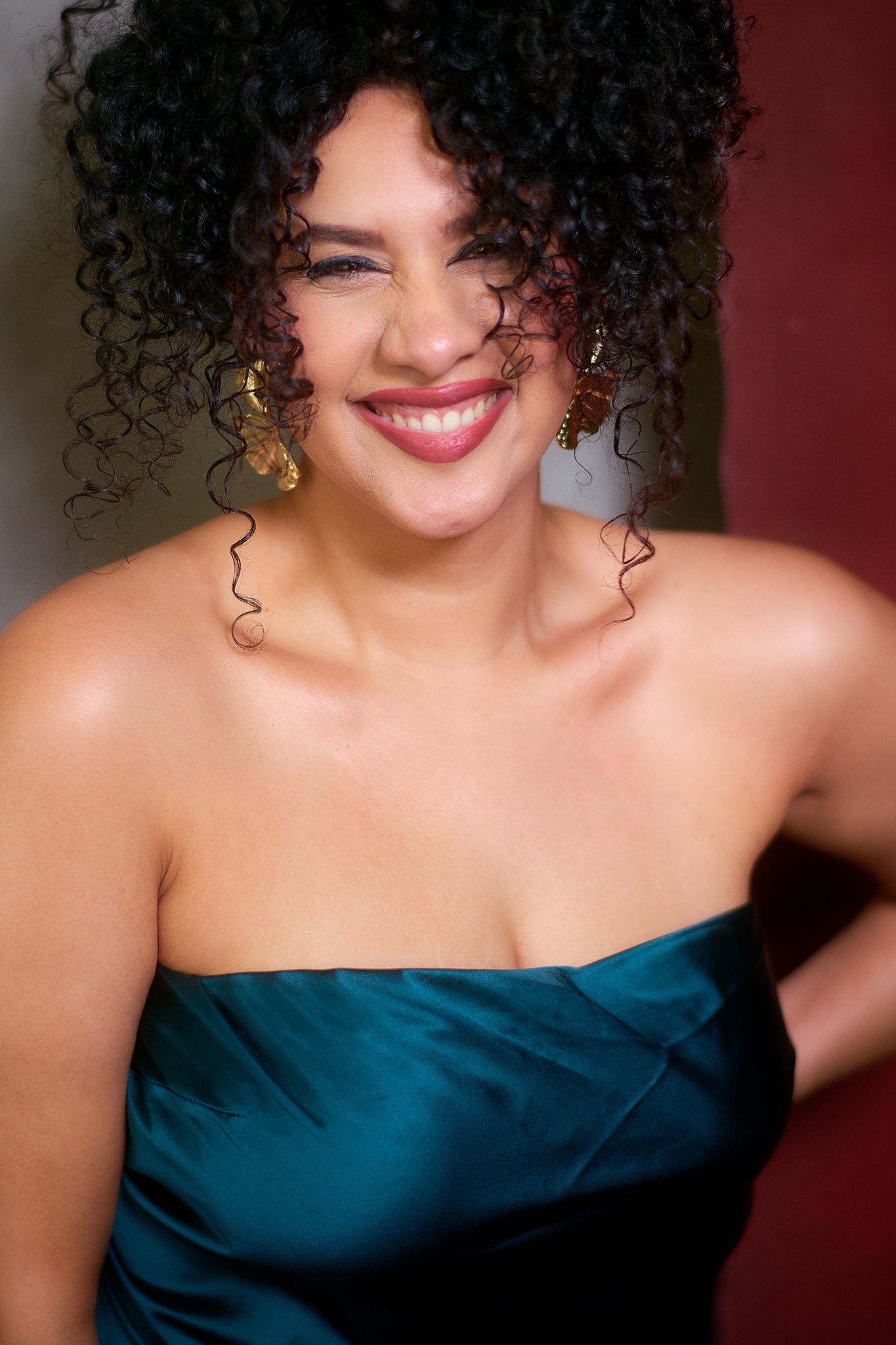 Smiling woman with curly hair wearing a teal strapless dress and gold earrings posing confidently.