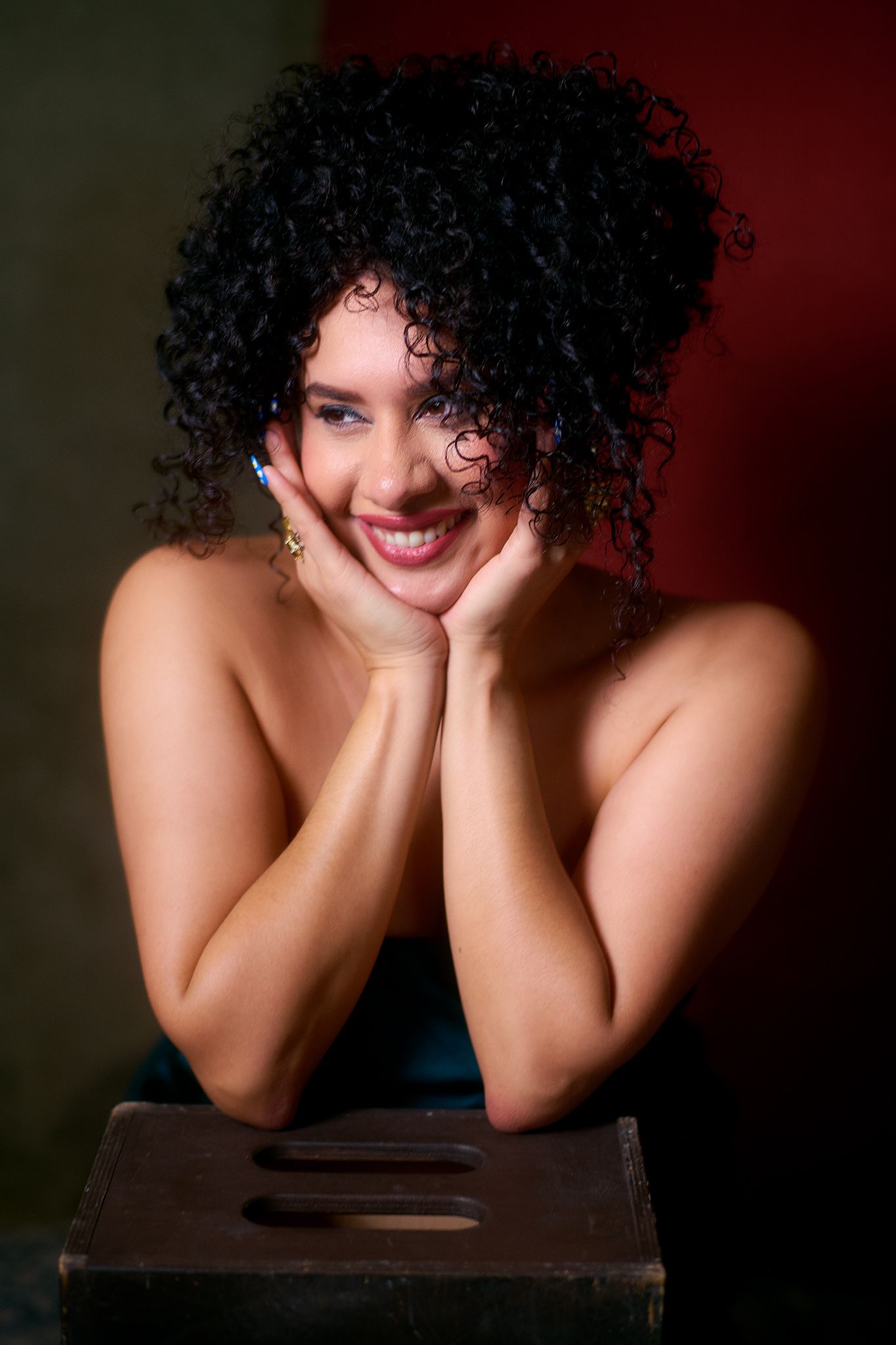 Smiling woman with curly black hair resting her face in her hands, posing against a colorful dark background.